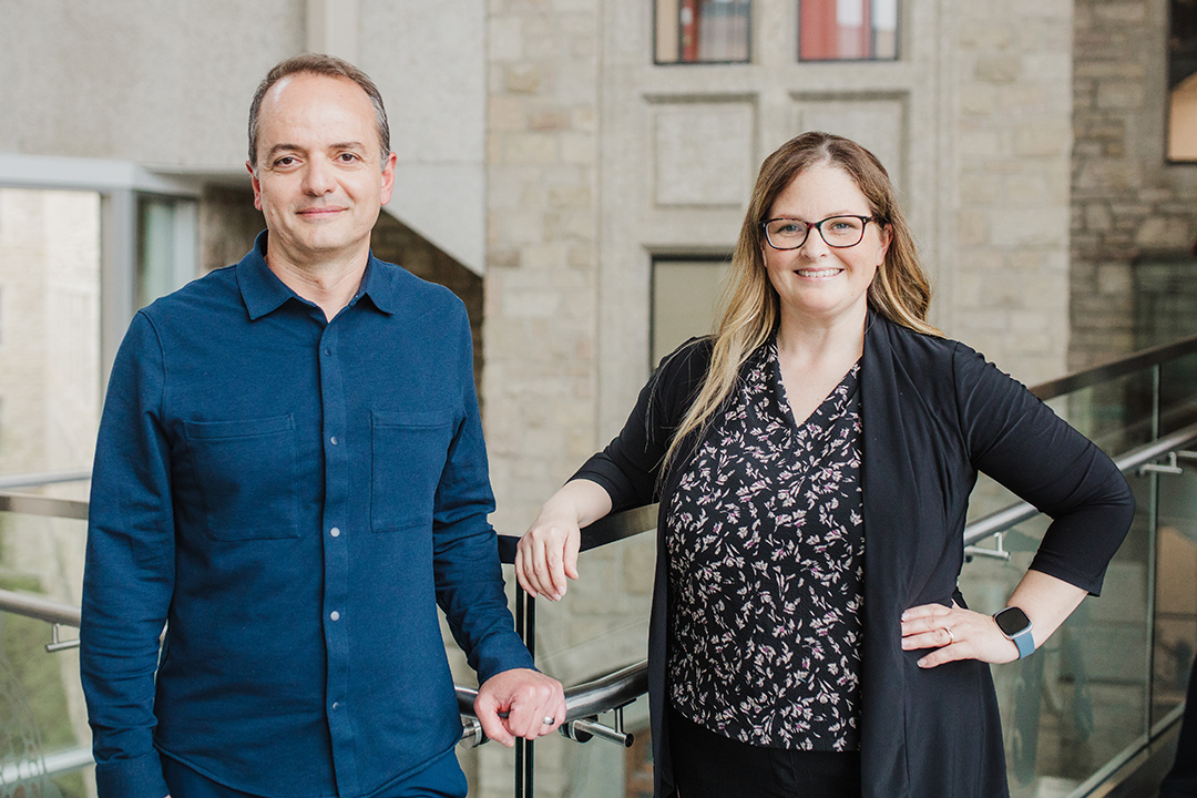 Man and woman standing in front of a railing with a stone facade behind them.